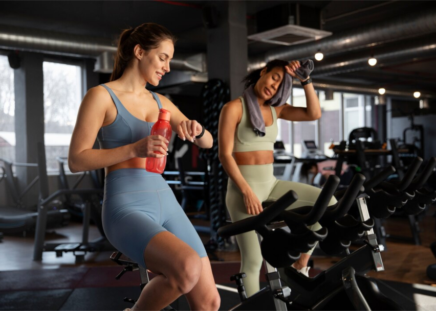 Спортивный зал в котором две девушки занимаются на велотренажёрах.