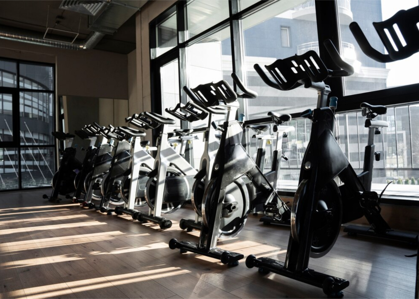 Велотренажёры в спортивном просторном зале, имеющий на заднем фоне широкие оконные рамы.